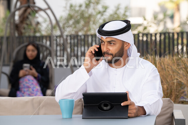اجراء مكالمة هاتفية ،الاستمتاع بالمشروبات الساخنه، استخدام جهاز التابلت لانجاز المشاريع في المقهى، اجراء مكالمة فيديو عبر الاجهزة التقنية الحديثة، العمل عن بعد، رجل عربي خليجي اماراتي يرتدي الكندورة والغترة البيضاء يجلس في كوفي شوب راقي بالامارات ، تجلس فى الخلف سيدة اماراتية تستخدم الجوال
