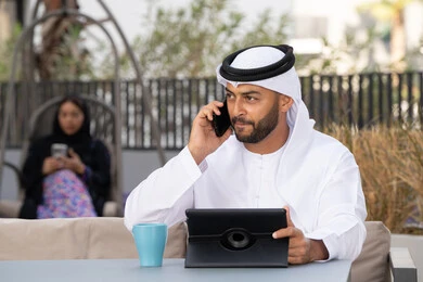 اجراء مكالمة هاتفية ،الاستمتاع بالمشروبات الساخنه، استخدام جهاز التابلت لانجاز المشاريع في المقهى، اجراء مكالمة فيديو عبر الاجهزة التقنية الحديثة، العمل عن بعد، رجل عربي خليجي اماراتي يرتدي الكندورة والغترة البيضاء يجلس في كوفي شوب راقي بالامارات ، تجلس فى الخلف سيدة اماراتية تستخدم الجوال