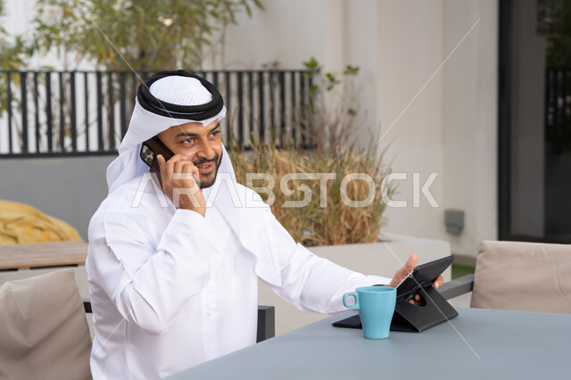 استخدام التكنولوجيا والتقنيات الحديثه، رجل عربي خليجي اماراتي مبتسم يرتدي الكندورة والغترة البيضاء يجلس في كوفي شوب راقي بالامارات ، العمل عن بعد، الاستمتاع بالمشروبات الساخنه، اجراء مكالمة هاتفية ،استخدام جهاز التابلت لانجاز المشاريع في المقهى
