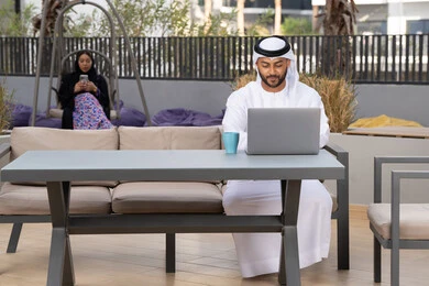 Working remotely, using a laptop to complete projects in a cafe, making a video call via modern technical devices, an Arab Gulf Emirati man wearing a white kandura and ghutra sitting in an upscale coffee shop in the Emirates, an Emirati woman sitting in the back using a mobile phone