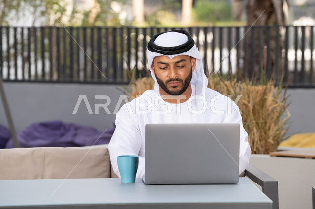 الاستمتاع بالمشروبات الساخنه ، العمل عن بعد،استخدام جهاز الحاسوب المحمول لانجاز المشاريع في المقهى، اجراء مكالمة فيديو عبر الاجهزة التقنية الحديثة، رجل عربي خليجي اماراتي يرتدي الكندورة والغترة البيضاء يجلس في كوفي شوب راقي بالامارات