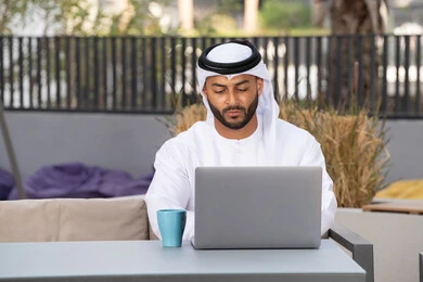 الاستمتاع بالمشروبات الساخنه ، العمل عن بعد،استخدام جهاز الحاسوب المحمول لانجاز المشاريع في المقهى، اجراء مكالمة فيديو عبر الاجهزة التقنية الحديثة، رجل عربي خليجي اماراتي يرتدي الكندورة والغترة البيضاء يجلس في كوفي شوب راقي بالامارات