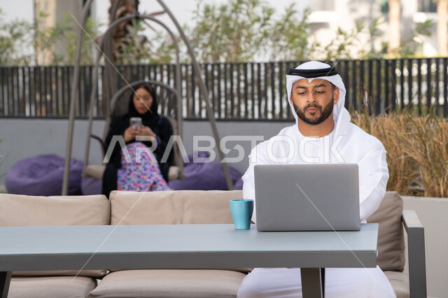Using a laptop to complete projects in a cafe, making a video call via modern technical devices, working remotely, a smiling Emirati Gulf Arab man wearing a white kandura and ghutra sitting in an upscale coffee shop in the Emirates, an Emirati woman sitting in the back using a mobile phone