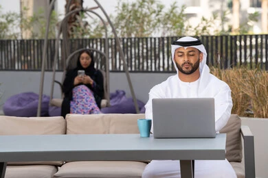 استخدام جهاز الحاسوب المحمول لانجاز المشاريع في المقهى، اجراء مكالمة فيديو عبر الاجهزة التقنية الحديثة، العمل عن بعد، رجل عربي خليجي اماراتي مبتسم يرتدي الكندورة والغترة البيضاء يجلس في كوفي شوب راقي بالامارات ، تجلس فى الخلف سيدة اماراتية تستخدم الجوال