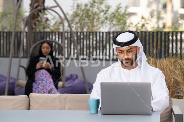 اجراء مكالمة فيديو عبر الاجهزة التقنية الحديثة، استخدام جهاز الحاسوب المحمول لانجاز المشاريع في المقهى، العمل عن بعد، رجل عربي خليجي اماراتي مبتسم يرتدي الكندورة والغترة البيضاء يجلس في كوفي شوب راقي بالامارات ، تجلس فى الخلف سيدة اماراتية تستخدم الجوال