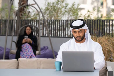 اجراء مكالمة فيديو عبر الاجهزة التقنية الحديثة، استخدام جهاز الحاسوب المحمول لانجاز المشاريع في المقهى، العمل عن بعد، رجل عربي خليجي اماراتي مبتسم يرتدي الكندورة والغترة البيضاء يجلس في كوفي شوب راقي بالامارات ، تجلس فى الخلف سيدة اماراتية تستخدم الجوال