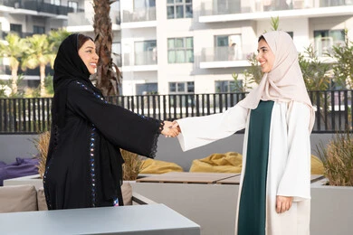 A relaxing weekend session, two Saudi women shaking hands with feelings of intimacy and love, spending a pleasant time with friends, two Arab Gulf Emirati friends sitting at a table in a luxury coffee shop in the Emirates