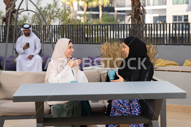 كوفي شوب راقي بالامارات، قضاء وقت ممتع مع الأصدقاء،  الاستمتاع بالمشروبات الساخنه ،رجل اماراتي فى الخلف يقوم باستخدم الجوال ،  تعابير السعادة والسرور بلمة الرفاق، تطوير العلاقات الإجتماعية،امرأتين عربيتين خليجيتين اماراتيتين تجلسان على طاولة في المقهى