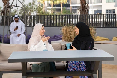 Upscale coffee shop in the Emirates, having a good time with friends, enjoying hot drinks, an Emirati man in the background using his mobile phone, expressions of happiness and pleasure at a gathering of friends, developing social relations, two Arab Gulf Emirati women sitting at a table in the cafe