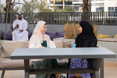 Having a good time with friends, enjoying hot drinks, an Emirati man in the background using his mobile phone, a high-end coffee shop in the Emirates, expressions of happiness and pleasure at a gathering of friends, developing social relations, two Arab Gulf Emirati women sitting at a table in a cafe