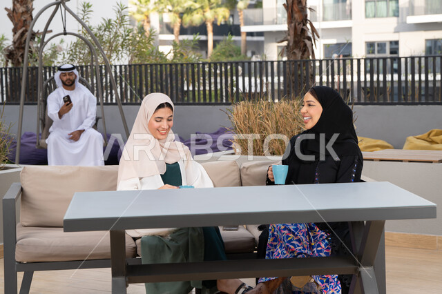 الاستمتاع بالمشروبات الساخنه ،رجل اماراتي فى الخلف يقوم باستخدم الجوال ، كوفي شوب راقي بالامارات، تعابير السعادة والسرور بلمة الرفاق، تطوير العلاقات الإجتماعية، قضاء وقت ممتع مع الأصدقاء، امرأتين عربيتين خليجيتين اماراتيتين تجلسان على طاولة في المقهى