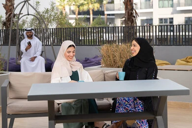 Enjoying hot drinks, Emirati man in the background using mobile phone, upscale coffee shop in the Emirates, expressions of happiness and pleasure with a gathering of friends, developing social relations, spending a good time with friends, two Emirati Gulf Arab women sitting at a table in the cafe