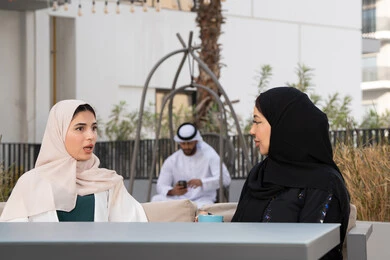 Emirati man in the background using mobile phone, upscale coffee shop in the Emirates, expressions of happiness and pleasure with a gathering of friends, developing social relations, spending a good time with friends, enjoying hot drinks, two Arab Gulf Emirati women sitting at a table in the cafe