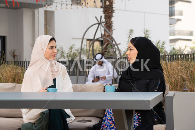 كوفي شوب راقي بالامارات، تعابير السعادة والسرور بلمة الرفاق، تطوير العلاقات الإجتماعية، قضاء وقت ممتع مع الأصدقاء، الاستمتاع بالمشروبات الساخنه ، رجل اماراتي فى الخلف يقوم باستخدم الجوال ، امرأتين عربيتين خليجيتين اماراتيتين تجلسان على طاولة في المقهى