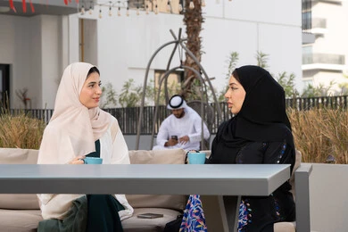 كوفي شوب راقي بالامارات، تعابير السعادة والسرور بلمة الرفاق، تطوير العلاقات الإجتماعية، قضاء وقت ممتع مع الأصدقاء، الاستمتاع بالمشروبات الساخنه ، رجل اماراتي فى الخلف يقوم باستخدم الجوال ، امرأتين عربيتين خليجيتين اماراتيتين تجلسان على طاولة في المقهى