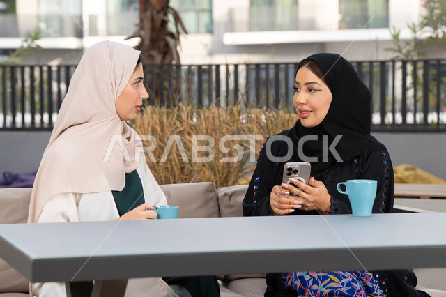تعابير السعادة والسرور بلمة الرفاق، تطوير العلاقات الإجتماعية، قضاء وقت ممتع مع الأصدقاء، الاستمتاع بالمشروبات الساخنه ، سيدة سعودية تقوم باستخدم الجوال ، كوفي شوب راقي بالامارات، امرأتين عربيتين خليجيتين اماراتيتين تجلسان على طاولة في المقهى