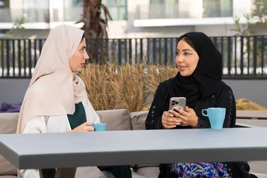 تعابير السعادة والسرور بلمة الرفاق، تطوير العلاقات الإجتماعية، قضاء وقت ممتع مع الأصدقاء، الاستمتاع بالمشروبات الساخنه ، سيدة سعودية تقوم باستخدم الجوال ، كوفي شوب راقي بالامارات، امرأتين عربيتين خليجيتين اماراتيتين تجلسان على طاولة في المقهى