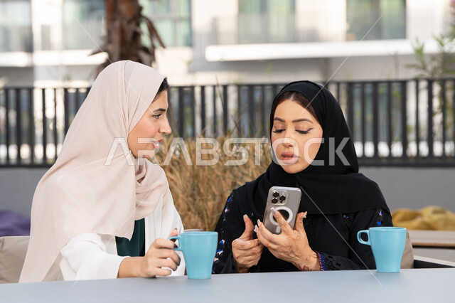 قضاء وقت ممتع مع الأصدقاء، الاستمتاع بالمشروبات الساخنه ، سيدة سعودية تقوم باستخدم الجوال ، كوفي شوب راقي بالامارات، تعابير السعادة والسرور بلمة الرفاق، امرأتين عربيتين خليجيتين اماراتيتين تجلسان على طاولة في المقهى ، تطوير العلاقات الإجتماعية