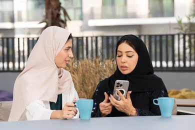 قضاء وقت ممتع مع الأصدقاء، الاستمتاع بالمشروبات الساخنه ، سيدة سعودية تقوم باستخدم الجوال ، كوفي شوب راقي بالامارات، تعابير السعادة والسرور بلمة الرفاق، امرأتين عربيتين خليجيتين اماراتيتين تجلسان على طاولة في المقهى ، تطوير العلاقات الإجتماعية