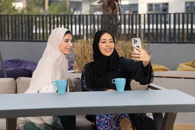 Saudi woman taking selfies, upscale coffee shop in UAE, expressions of happiness and pleasure with friends gathering, spending quality time with friends, enjoying hot drinks, two Emirati Gulf Arab women sitting at a table in a cafe, developing social relations
