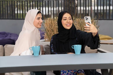 Upscale coffee shop in the Emirates, expressions of happiness and pleasure with a gathering of friends, spending a pleasant time with friends, enjoying hot drinks, two Emirati Gulf Arab women sitting at a table in the cafe, a Saudi woman taking souvenir selfies, developing social relations