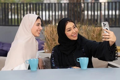 Expressions of happiness and pleasure, gathering with friends, having a good time with friends, enjoying hot drinks, two Emirati Gulf Arab women sitting at a table in a cafe, a Saudi woman taking souvenir selfies, developing social relations, a high-end coffee shop in the Emirates