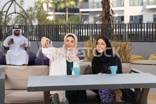 الاستمتاع بالمشروبات الساخنه ، امرأتين عربيتين خليجيتين اماراتيتين تجلسان على طاولة في المقهى وبخلفهم رجل اماراتي يستخدم الجوال ، التقاط صور سيلفي تذكارية ، تطوير العلاقات الإجتماعية، تعابير السعادة والسرور بلمة الرفاق، كوفي شوب راقي بالامارات، قضاء وقت ممتع مع الأصدقاء.