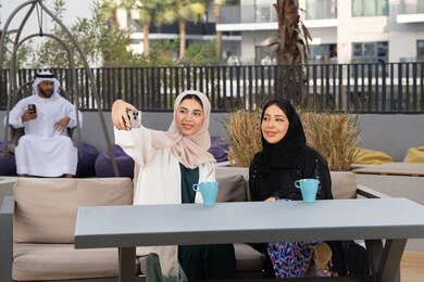 Enjoying hot drinks, two Emirati Gulf Arab women sitting at a table in a cafe with an Emirati man behind them using his mobile phone, taking souvenir selfies, developing social relationships, expressions of happiness and pleasure at a gathering of friends, an upscale coffee shop in the Emirates, spending a pleasant time with friends.