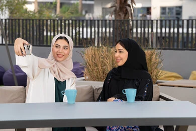 A Saudi woman taking selfies, enjoying a hot drink, two Emirati Gulf Arab women sitting at a table in a cafe, developing social relationships, expressions of happiness and pleasure at a gathering of friends, a high-end coffee shop in the Emirates, spending quality time with friends.