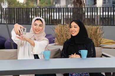 Two Emirati Gulf Arab women sitting at a table in a cafe, a Saudi woman taking selfies, enjoying hot drinks, developing social relationships, expressions of happiness and pleasure at a gathering of friends, an upscale coffee shop in the Emirates, spending quality time with friends.