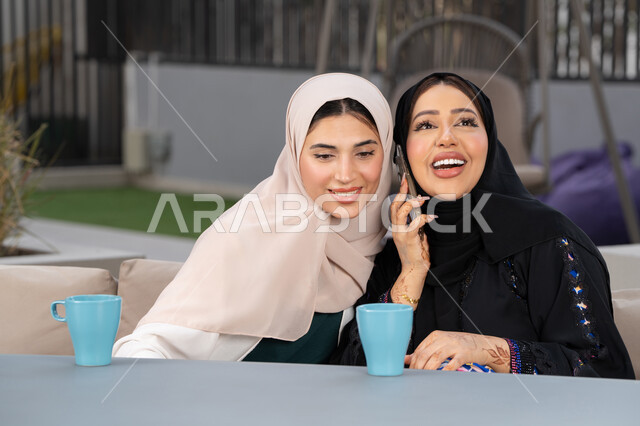 الاستمتاع بالمشروبات الساخنه ، امرأتين عربيتين خليجيتين اماراتيتين تجلسان على طاولة في المقهى، تطوير العلاقات الإجتماعية، سيدة سعودية تقوم باجراء مكالمه هاتفية عبر الجوال ، تعابير السعادة والسرور بلمة الرفاق، كوفي شوب راقي بالامارات، قضاء وقت ممتع مع الأصدقاء.