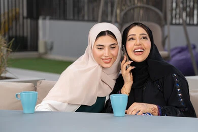 Enjoying hot drinks, two Emirati Gulf Arab women sitting at a table in a cafe, developing social relationships, a Saudi woman making a phone call on her mobile phone, expressions of happiness and pleasure at a gathering of friends, a classy coffee shop in the Emirates, spending quality time with friends.