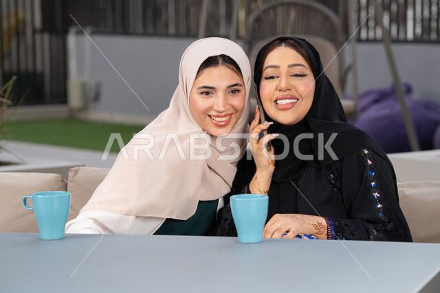 امرأتين عربيتين خليجيتين اماراتيتين تجلسان على طاولة في المقهى، تطوير العلاقات الإجتماعية، سيدة سعودية تقوم باجراء مكالمه هاتفية عبر الجوال ، تعابير السعادة والسرور بلمة الرفاق، كوفي شوب راقي بالامارات، قضاء وقت ممتع مع الأصدقاء،  الاستمتاع بالمشروبات الساخنه .