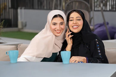 Two Emirati Gulf Arab women sitting at a table in a cafe, developing social relationships, a Saudi woman making a phone call on her mobile phone, expressions of happiness and pleasure at a gathering of friends, an upscale coffee shop in the Emirates, spending a pleasant time with friends, enjoying hot drinks.
