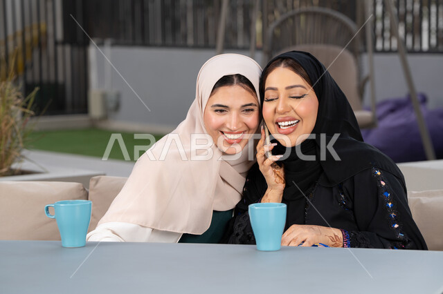 تطوير العلاقات الإجتماعية، سيدة سعودية تقوم باجراء مكالمه هاتفية عبر الجوال ، تعابير السعادة والسرور بلمة الرفاق، كوفي شوب راقي بالامارات، قضاء وقت ممتع مع الأصدقاء، امرأتين عربيتين خليجيتين اماراتيتين تجلسان على طاولة في المقهى، الاستمتاع بالمشروبات الساخنه .