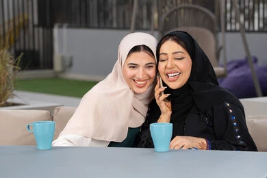 Developing social relationships, a Saudi woman making a phone call, expressions of happiness and pleasure at a gathering of friends, a high-end coffee shop in the Emirates, spending quality time with friends, two Emirati Gulf Arab women sitting at a table in a cafe, enjoying hot drinks.