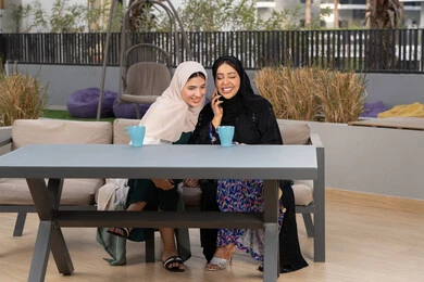 A Saudi woman making a phone call on her mobile phone, expressions of happiness and pleasure at a gathering of friends, a high-end coffee shop in the Emirates, spending a pleasant time with friends, two Arab Gulf Emirati women sitting at a table in a cafe, enjoying hot drinks, developing social relations.