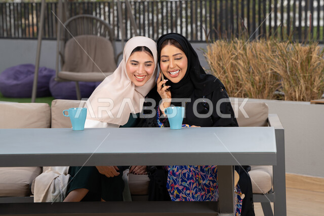 Expressions of happiness and pleasure at a gathering of friends, a high-end coffee shop in the Emirates, spending a pleasant time with friends, two Emirati Gulf Arab women sitting at a table in a cafe, enjoying hot drinks, a Saudi woman making a phone call on her mobile phone, developing social relations.