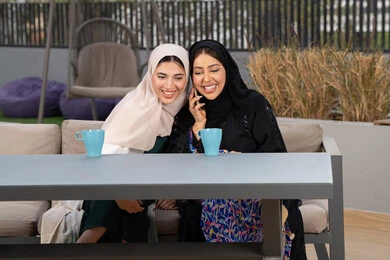 تعابير السعادة والسرور بلمة الرفاق، كوفي شوب راقي بالامارات، قضاء وقت ممتع مع الأصدقاء، امرأتين عربيتين خليجيتين اماراتيتين تجلسان على طاولة في المقهى، الاستمتاع بالمشروبات الساخنه ،  سيدة سعودية تقوم باجراء مكالمه هاتفية عبر الجوال ، تطوير العلاقات الإجتماعية.