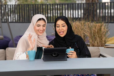 Having a good time with friends, two Emirati Gulf Arab women sitting at a table in a cafe, enjoying hot drinks, expressions of happiness and pleasure at the gathering of friends, two Saudi friends making a video call via tablet, a high-end coffee shop in the Emirates, developing social relations.