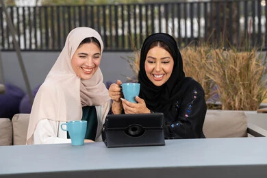 امرأتين عربيتين خليجيتين اماراتيتين تجلسان على طاولة في المقهى، الاستمتاع بالمشروبات الساخنه ، تعابير السعادة والسرور بلمة الرفاق، صديقتين سعوديتين يقومان باجراء مكالمه فيديو عبر التابلت ، كوفي شوب راقي بالامارات، التسامر والدردشة وقضاء وقت ممتع مع الأصدقاء، تطوير العلاقات الإجتماعية.