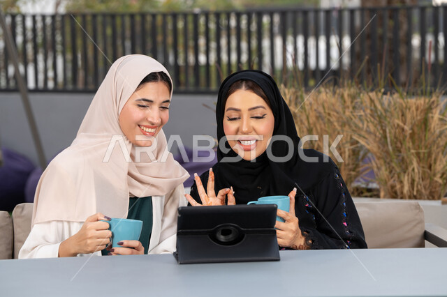 Enjoying hot drinks, expressions of happiness and pleasure with friends, two Saudi friends making a video call via tablet, a high-end coffee shop in the Emirates, chatting and having a good time with friends, developing social relationships, two Arab Gulf Emirati women sitting at a table in a cafe.