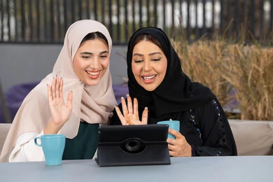 Expressions of happiness and pleasure at a gathering of friends, two Saudi friends making a video call via tablet, a high-end coffee shop in the Emirates, chatting and having a good time with friends, developing social relationships, two Arab Gulf Emirati women sitting at a table in a cafe, enjoying hot drinks.