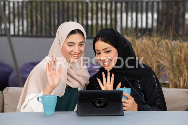 Two Saudi friends making a video call via tablet, a high-end coffee shop in the Emirates, chatting and having a good time with friends, developing social relationships, expressions of happiness and pleasure at a gathering of friends, two Arab Gulf Emirati women sitting at a table in a cafe, enjoying hot drinks.