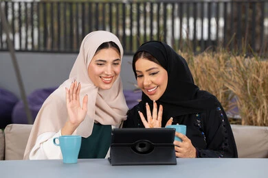 صديقتين سعوديتين يقومان باجراء مكالمه فيديو عبر التابلت ، كوفي شوب راقي بالامارات، التسامر والدردشة وقضاء وقت ممتع مع الأصدقاء، تطوير العلاقات الإجتماعية، تعابير السعادة والسرور بلمة الرفاق، امرأتين عربيتين خليجيتين اماراتيتين تجلسان على طاولة في المقهى، الاستمتاع بالمشروبات الساخنه .