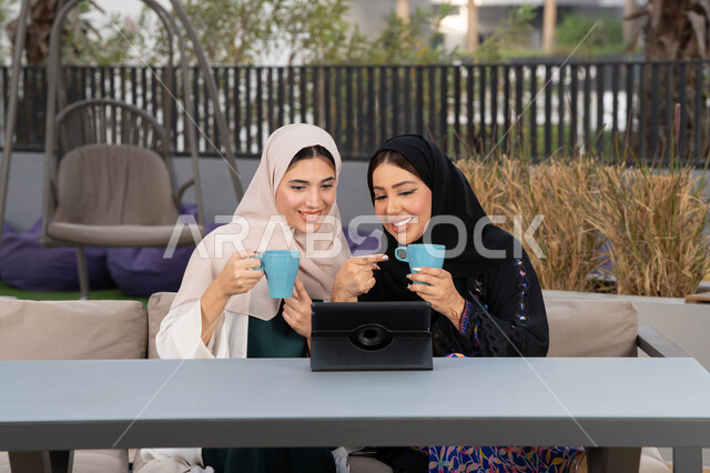 كوفي شوب راقي بالامارات، التسامر والدردشة وقضاء وقت ممتع مع الأصدقاء، تطوير العلاقات الإجتماعية، تعابير السعادة والسرور بلمة الرفاق، امرأتين عربيتين خليجيتين اماراتيتين تجلسان على طاولة في المقهى، صديقتين سعوديتين يقومان باجراء مكالمه فيديو عبر التابلت ، الاستمتاع بالمشروبات الساخنه .