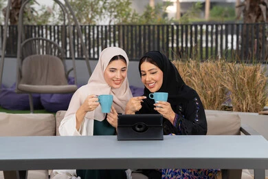 كوفي شوب راقي بالامارات، التسامر والدردشة وقضاء وقت ممتع مع الأصدقاء، تطوير العلاقات الإجتماعية، تعابير السعادة والسرور بلمة الرفاق، امرأتين عربيتين خليجيتين اماراتيتين تجلسان على طاولة في المقهى، صديقتين سعوديتين يقومان باجراء مكالمه فيديو عبر التابلت ، الاستمتاع بالمشروبات الساخنه .