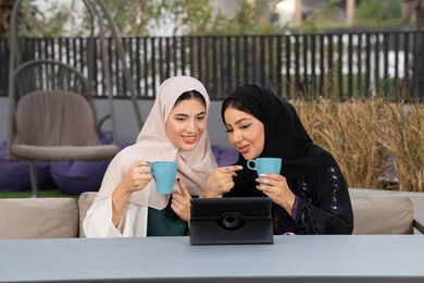 التسامر والدردشة وقضاء وقت ممتع مع الأصدقاء، تطوير العلاقات الإجتماعية،كوفي شوب راقي بالامارات، تعابير السعادة والسرور بلمة الرفاق، امرأتين عربيتين خليجيتين اماراتيتين تجلسان على طاولة في المقهى، صديقتين سعوديتين يقومان باجراء مكالمه فيديو عبر التابلت ، الاستمتاع بالمشروبات الساخنه .