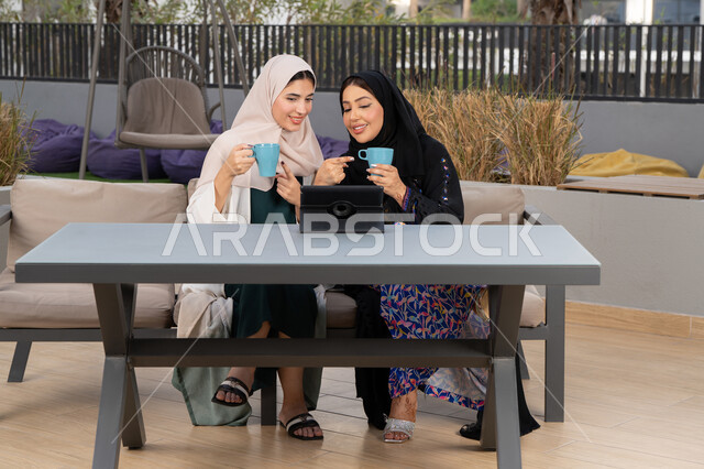 Developing social relationships, chatting and having a good time with friends, a classy coffee shop in the Emirates, expressions of happiness and pleasure at a gathering of friends, two Emirati Gulf Arab women sitting at a table in a cafe, two Saudi friends making a video call via tablet, enjoying hot drinks.