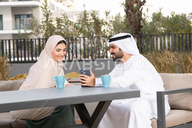 Gestures of pleasure and happiness, spending honeymoon, enjoying hot drinks, an Arab man taking a selfie with his wife, the concept of affection and intimacy, spending a pleasant time, a smiling Emirati Gulf Arab couple sitting in a cafe